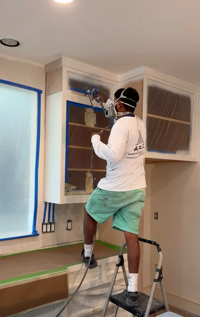 Painter spray-applying white finish to kitchen cabinet doors with blue painter's tape masking