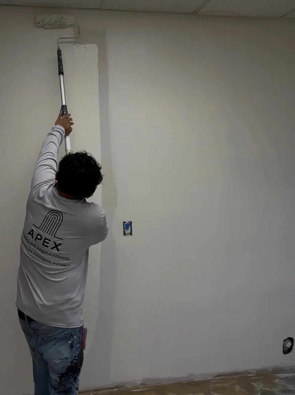 Worker applying white paint to interior drywall wall with roller and extension pole